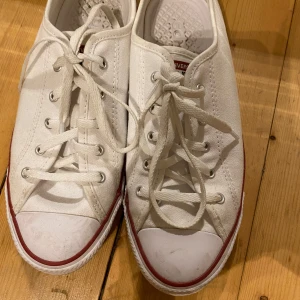 Vita Converse All Star sneakers - Säljer ett par klassiska vita Converse All Star sneakers med snörning. Skorna har en röd rand längs sulan och är perfekta för en avslappnad stil.
