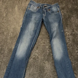 Blå jeans från True Religion - Snygga blå jeans från True Religion med bootcut-stil. De har en klassisk femficksdesign och tydliga sömmar som ger en cool look. Perfekta för en avslappnad stil. Har ett hål i bakfickan men går absolut att fixa genom att sy !! Det är storlek L på jeansen men funkar som M också !! 