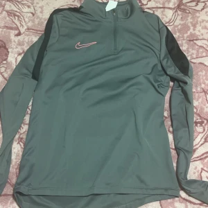 Grå långärmad tröja från Nike - Säljer en grå långärmad tröja från Nike med Dri-FIT-teknologi. Tröjan har en halv dragkedja och ett rött Nike-märke på bröstet. Perfekt för träning eller vardagsbruk.