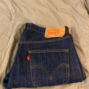 Levi's 501 jeans - Klassiska Levi's 501 jeans i mörkblå denim. De har en rak passform och är tillverkade i slitstarkt jeansmaterial. Perfekta för en avslappnad stil med den ikoniska Levi's-lappen på baksidan och den röda etiketten vid fickan.