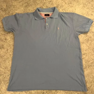 Blå pikétröja från Ralph Lauren - Säljer en klassisk blå pikétröja från Ralph Lauren med korta ärmar och en broderad logotyp på bröstet. Tröjan har en krage och knappar framtill. Perfekt för en stilren look.