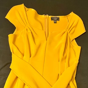 Gul klänning från Lipsy London - Snygg gul klänning från Lipsy London med en elegant knytning i midjan. Klänningen har en v-ringad design och korta ärmar, perfekt för en stilfull look. Den har en dragkedja i ryggen för enkel påklädning.