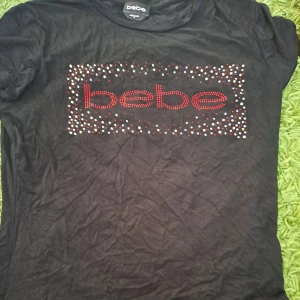 Svart t-shirt från Bebe med paljetter - Snygg svart t-shirt från Bebe med röd logga och glittrande paljetter på framsidan. Perfekt för att ge din outfit lite extra bling. Kortärmad och bekväm passform. Y2k style 