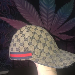 Mönstrad keps från Gucci - Snygg keps från Gucci med ett klassiskt mönster. Kepsen har en röd och blå randdetalj på sidan och är tillverkad i ett slitstarkt material. Perfekt för att ge din outfit en lyxig touch.