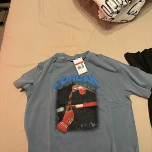 Grå Jordan t-shirt med tryck - Säljer en grå t-shirt från Jordan med ett coolt tryck av en basketspelare i aktion. T-shirten har korta ärmar och en klassisk passform. Perfekt för sportiga tillfällen eller som en stilren vardagslook.