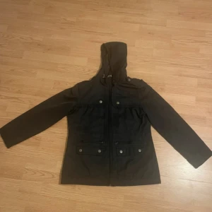 Svart jacka med huva från Reitmans - Säljer en svart jacka med huva från Reitmans. Jackan har långa ärmar och praktiska fickor med knappar framtill. Perfekt för kyligare dagar med sin stilrena design.