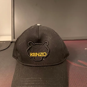 Svart keps från Kenzo - Snygg svart keps från Kenzo med broderad logotyp i gult framtill och baktill. Kepsen har en justerbar rem för perfekt passform. Perfekt för att ge din outfit en stilren touch.