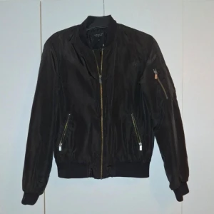 Svart bomberjacka med dragkedjor - Snygg svart bomberjacka med dragkedjor fram och på ärmen. Jackan har en klassisk bomberstil med ribbade muddar vid ärmslut och nederkant. Perfekt för en cool och avslappnad look.