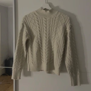 Vit stickad tröja med mönster - Säljer en mysig vit stickad tröja med ett snyggt kabelstickat mönster. Tröjan har långa ärmar och en hög krage som håller dig varm under kyliga dagar. Perfekt för höst och vinter!