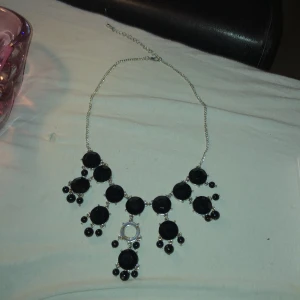 Svart halsband med stora stenar - Elegant halsband med stora svarta stenar och små pärldetaljer. Kedjan är i silverfärg och justerbar för perfekt passform. Perfekt för att ge en stilren touch till din outfit.