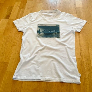 Vit t-shirt från Pepe Jeans London - Säljer en vit t-shirt från Pepe Jeans London med ett tryck av roddare och texten 'Portobello Rd Mkt Rowing Club 1973'. T-shirten har en slim passform och är kortärmad. Perfekt för en avslappnad stil.