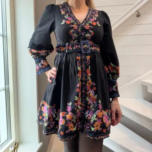 Svart blommig klänning med bälte från By Malina - Charmig svart klänning med färgglada blommor och ett matchande bälte i midjan. Klänningen har långa ärmar och en v-ringad hals. Perfekt för en bohemisk look med sina livfulla blommönster.