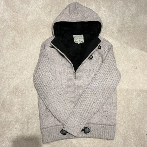 Fodrad Cardigan Jacka - Säljer en grå stickad jacka med huva och dragkedja. Jackan har snygga knapplås framtill och ribbade detaljer på ärmar och nederkant. Perfekt för kyliga dagar! Vid minsta fråga eller fundering hör av er🤝