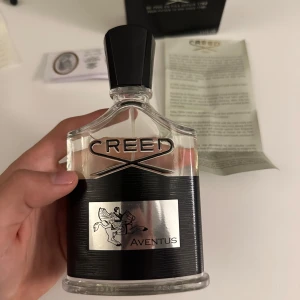 Creed Aventus Parfym 100ml - Säljer en exklusiv Creed Aventus parfym i en elegant glasflaska med svart och silver detaljer. Flaskan rymmer 100 ml och har en stilren svart kork. Aventus är känd för sin unika doftprofil med inslag av frukt och trä.