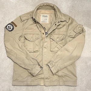 Ralph Lauren - Field Jacket - Säljer en beige jacka från Ralph Lauren med coola patchar på ärmen. Jackan har flera fickor med knappar och en dragkedja framtill. Perfekt för en avslappnad stil. Vid minsta fråga eller fundering hör av er🤝