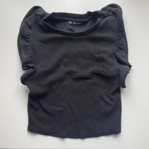 Svart ribbad topp från Zara - Säljer en stilren svart ribbad topp från Zara. Toppen har korta volangärmar och en rund halsringning. Perfekt för en enkel och snygg outfit. Den är i M men passar XS och S också