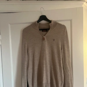 Beige merinoulltröja från Morris - Säljer en stilren beige tröja från Morris i 100% merinoull. Tröjan har en dragkedja vid halsen och ett diskret broderat emblem på bröstet. Perfekt för kyligare dagar.