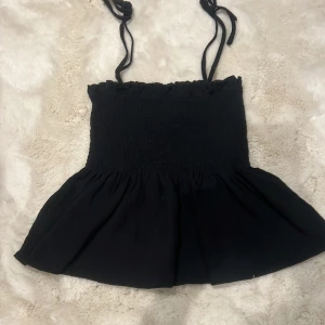 Svart axelbandslös topp - Snygg svart axelbandslös topp med smockdetaljer och knytband. Perfekt för varma sommardagar eller en kväll ute. Den har en luftig passform och en lätt volang vid nederkanten.