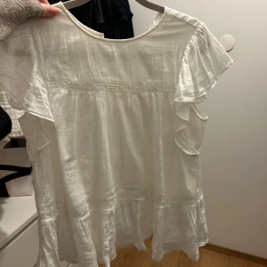 Vit blus med volangdetaljer - Charmig vit blus med volangdetaljer på ärmarna och nederkanten. 