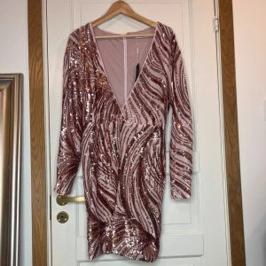 Rosa paljettklänning från NLY One - Säljer en glittrig rosa klänning från NLY One med paljetter i ett vågigt mönster. Klänningen har en djup v-ringning och långa ärmar, 