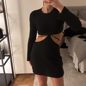 Svart klänning med cut-outs - Trendig svart klänning med cut-outs i midjan och långärmad design. Perfekt för en kväll ute med vänner. Klänningen har en tight passform och är kort i modellen.
