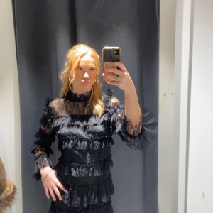 Svart spetsklänning från By Malina - Elegant svart klänning från By Malina i spets med volangdetaljer. Klänningen har långa ärmar och en hög hals, vilket ger en sofistikerad look. Perfekt för speciella tillfällen. Nypris 2800kr helt ny med prislapp kvar 