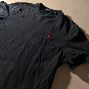Svart t-shirt från Ralph Lauren - Snygg svart t-shirt från Ralph Lauren med det klassiska röda broderade logotypen på bröstet. Perfekt för en stilren och avslappnad look. Tillverkad i mjukt material för extra komfort. Tröjan är i nyskick, pris kan självklart diskuteras vid snabb affär, mvh Eric
