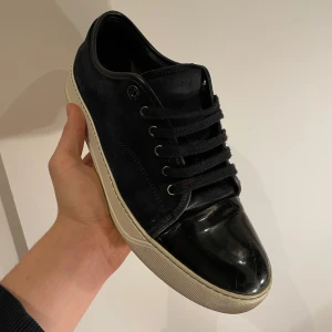 Svarta sneakers från Lanvin  - Snygga svarta sneakers med en kombination av mocka och lackdetaljer. Skorna har en klassisk snörning och en vit sula som ger en stilren kontrast. Perfekta för en trendig look.
