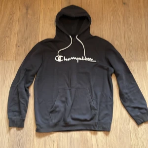 Svart hoodie från Champion - Säljer en svart hoodie från Champion med vit logga på bröstet. Den har en klassisk känguruficka och justerbar huva med vita snören. Perfekt för en avslappnad stil. Köptes för 350kr säljs för 250kr