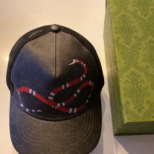 Svart Gucci keps med ormmotiv. Box ingår. - Snygg svart keps från Gucci med ett iögonfallande ormmotiv i rött, vitt och svart. Kepsen har ett nätmaterial baktill för extra ventilation och en justerbar rem för perfekt passform. Pris kan diskuteras lite.