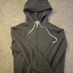 Grå hoodie från Polo Ralph Lauren - Säljer en stilren grå hoodie från Polo Ralph Lauren med dragkedja och vit snörning i huvan. Den har en liten grön logga på bröstet och är perfekt för en avslappnad look.
