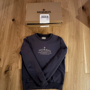 Mörkblå sweatshirt från Morris - Säljer en stilren mörkblå sweatshirt från Morris med texten 'Morris Gentlemen's Club' på bröstet. Tröjan har långa ärmar och ribbade muddar vid ärmslut och nederkant. Perfekt för en avslappnad stil.