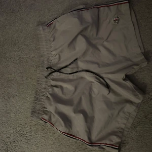 Beige shorts med snörning - Snygga beige shorts med elastisk midja och snörning för justerbar passform. De har en diskret logga på sidan och ränder i rött och svart längs kanterna. Perfekta för en avslappnad stil.