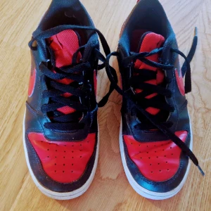 Svarta och röda Nike sneakers - Säljer ett par svarta och röda Nike sneakers med snörning. Skorna har en klassisk design med röd Nike-logga på sidan och perforerade detaljer på tån. Perfekta för en sportig look. Sula 23.5