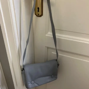 axelväska - Snygg ljusblå axelväska med enkel design och två knappar framtill. Perfekt för att bära dina nödvändigheter med stil. Justerbar axelrem för bekväm passform.