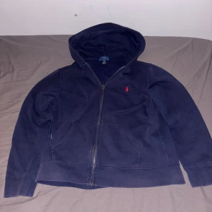 Mörkblå hoodie från Ralph Lauren - Säljer en snygg mörkblå hoodie från Ralph Lauren med dragkedja och den klassiska röda loggan på bröstet. Perfekt för en avslappnad stil. Den har en bekväm passform och huva för extra mysfaktor.