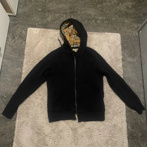 Svart huvtröja med rutig foder - Säljer en stilren svart huvtröja från Burberry med dragkedja och klassiskt rutigt foder i huvan. Tröjan har långa ärmar och ribbade muddar. Perfekt för en casual look. DEN HAR BLIVIT FÖR LITEN FÖR MIG SÅ DET ÄR DRF JAG SÄLJER. Kan gå ner i pris, max 200kr vid snabb affär.