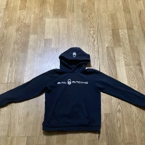 Svart hoodie från Sail Racing - Säljer en svart hoodie från Sail Racing med logga på bröstet. Den har en klassisk design med lång ärm och luva, perfekt för en avslappnad stil. Passar bra till både jeans och träningsbyxor.