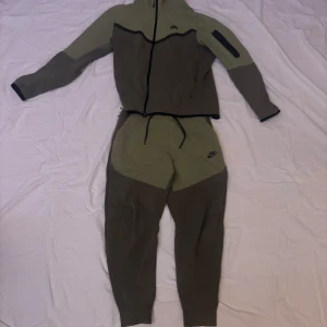 Grön Nike Tech - Snygg och bekväm grön träningsdräkt från Nike. Setet består av en huvtröja med dragkedja och matchande byxor. Perfekt för träning eller avslappnade dagar. OBS! Finns en liten fläck på vänstra byxbenet. Tröjan är L och byxorna är M. Om man inte vill ha båda kan man köpa dem för 300kr styck !