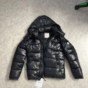 Svart pufferjacka från Moncler - Säljer en snygg svart pufferjacka från Moncler med huva och dragkedja. Jackan har en glansig finish och Moncler-logga på ärmen. Perfekt för kyliga dagar.