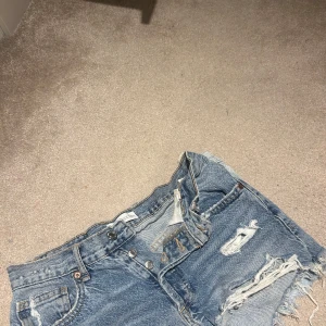 Blå jeansshorts med slitningar - Säljer ett par coola blå jeansshorts med slitningar. Perfekta för en avslappnad stil. De har en klassisk femficksdesign och knappgylf.