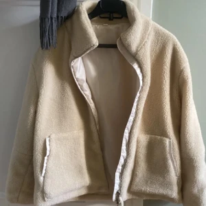 Beige teddyjacka med dragkedja - Mysig beige teddyjacka med dragkedja och två stora fickor framtill. Perfekt för kyliga dagar med sin mjuka och varma känsla. Jackan har en enkel design med hög krage.