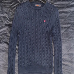 Mörkblå kabelstickad tröja med röd logga - Säljer en stilren mörkblå kabelstickad tröja med en liten röd logga på bröstet. Tröjan har långa ärmar och en rund halsringning. Perfekt för kyligare dagar. Modellen på bild 3 är cirka 190. Priset kan diskuteras!