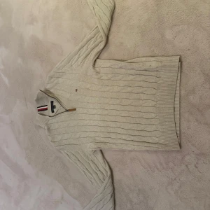Beige stickad tröja från Tommy Hilfiger - Säljer en stilren beige stickad tröja från Tommy Hilfiger med dragkedja och ribbade detaljer. Tröjan har ett klassiskt kabelstickat mönster och en liten logotyp på bröstet. Perfekt för kyligare dagar. St e lite stor så skulle nog säga att den passar s mer 