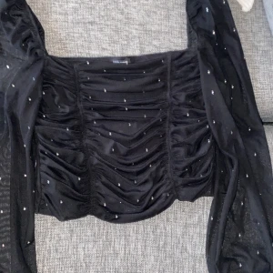 Svart glittrig topp  - Snygg svart topp från Bubbleroom med glittriga detaljer. Toppen har långa ärmar och en rynkad design framtill. Perfekt för en kväll ute eller en speciell tillställning.