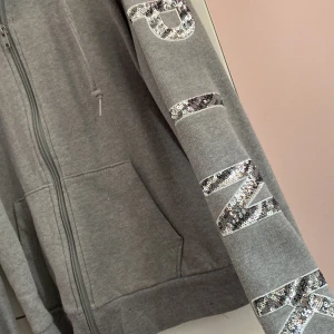 Grå zip up hoodie från Victoria's Secret PINK - Säljer en grå hoodie från Victoria's Secret PINK med glittriga bokstäver på ärmen. Den har dragkedja framtill och en mysig huva. Perfekt för en avslappnad stil. 🩶✨