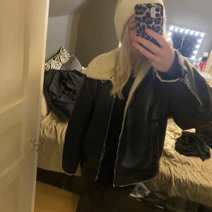 Svart skinnjacka med foder - Säljer en svart skinnjacka med mysigt vitt foder. Jackan har en dragkedja framtill och långa ärmar. Perfekt för kyligare dagar när du vill hålla stilen.
