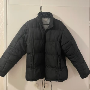 Svart pufferjacka från Diana Italia - Säljer en svart pufferjacka från Diana Italia i storlek S. Jackan har en hög krage och dragkedja framtill. Perfekt för kalla vinterdagar med sin pufferstil och varma fyllning. Den har en justerbar dragsko i nederkanten för extra skydd mot kyla. Använd men i mycket bra skick!