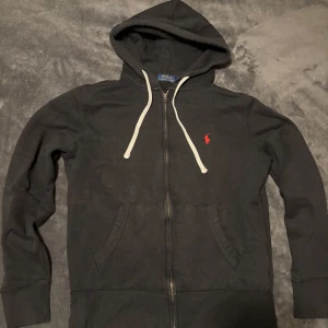 Ralph Lauren kofta - Säljer en stilren svart hoodie från Ralph Lauren med dragkedja och vit snörning. Den har en klassisk röd logga på bröstet och är perfekt för en avslappnad look. Storlek S.