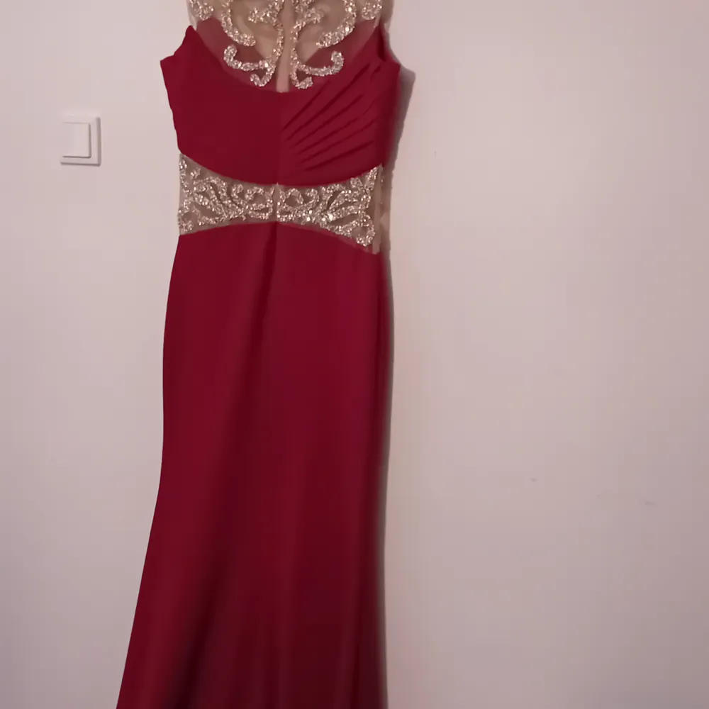 Elegant vinröd långklänning med vackra spetsdetaljer i guld. Klänningen har en figurnära passform och är ärmlös med en öppen rygg. Perfekt för festliga tillfällen som nyår eller bal. Materialet är mjukt och följsamt, vilket ger en lyxig känsla.. Mekot.